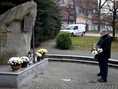 dwie osoby stoją przed pomnikiem - głazem; mężczyzna trzyma na rękach wiązankę biało czerwonych kwiatów