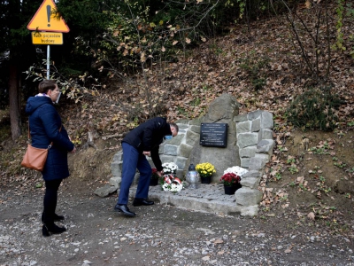 kobieta stoi za pochylonym mężczyzną który składa wiązankę pod pomnikiem z kamienia
