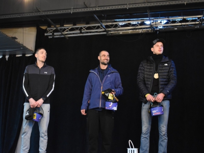 Trzech mężczyzn w strojach sportowych stoi na scenie. Pozują do zdjęcia z nagrodami.